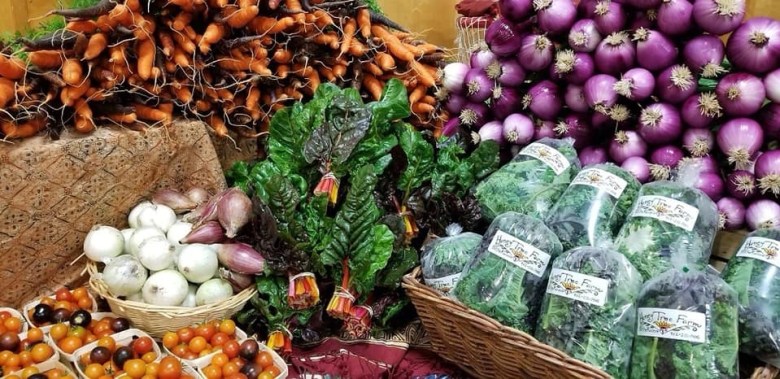 An image of a Farmer's Market table featuring carrots, onions, tomatoes and greens.