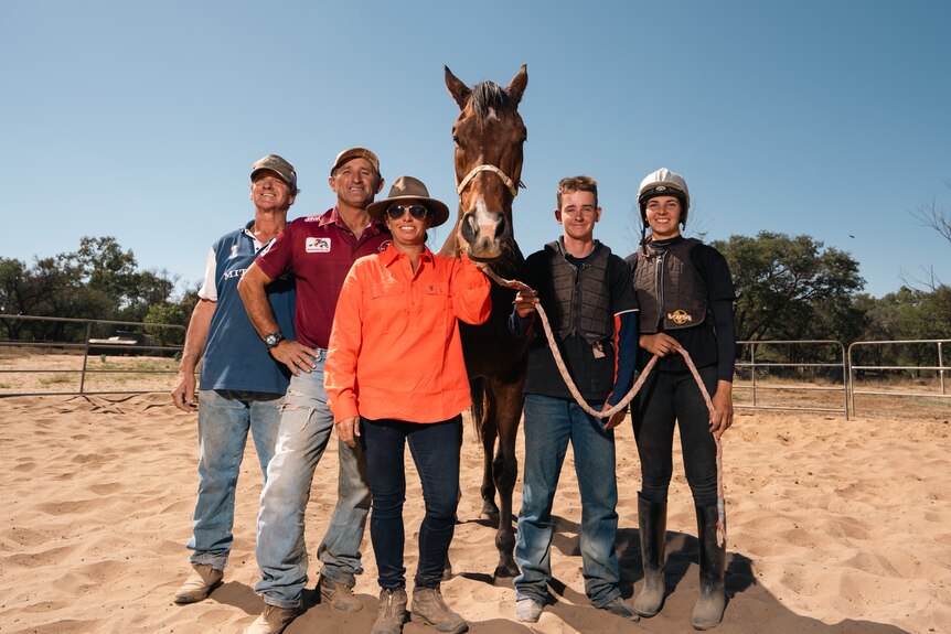 Five smiling people stand in a dusty paddock with a brown horse.