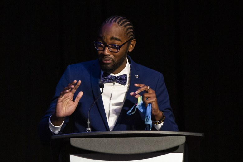 A Black man in a suit speaks at a podium, holding a blue mask in his hands.