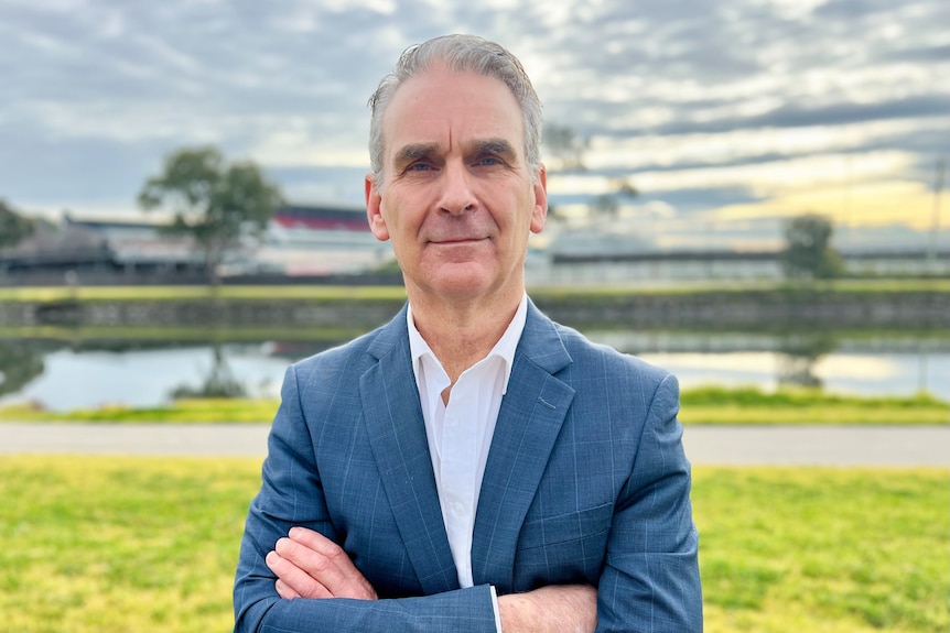 A man with grey hair stands in front of a camera with a racing track in the background