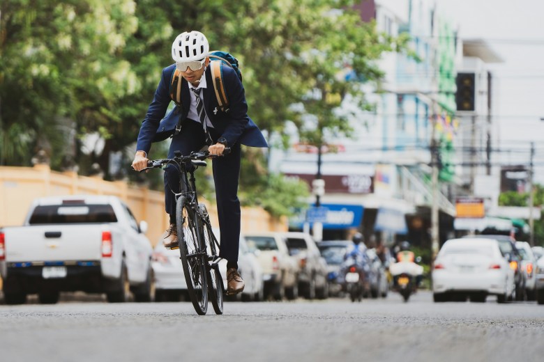 A man wearing a jacket and tie bikes down the middle of a street. He has a backpack, and wears a helmet and sunglasses