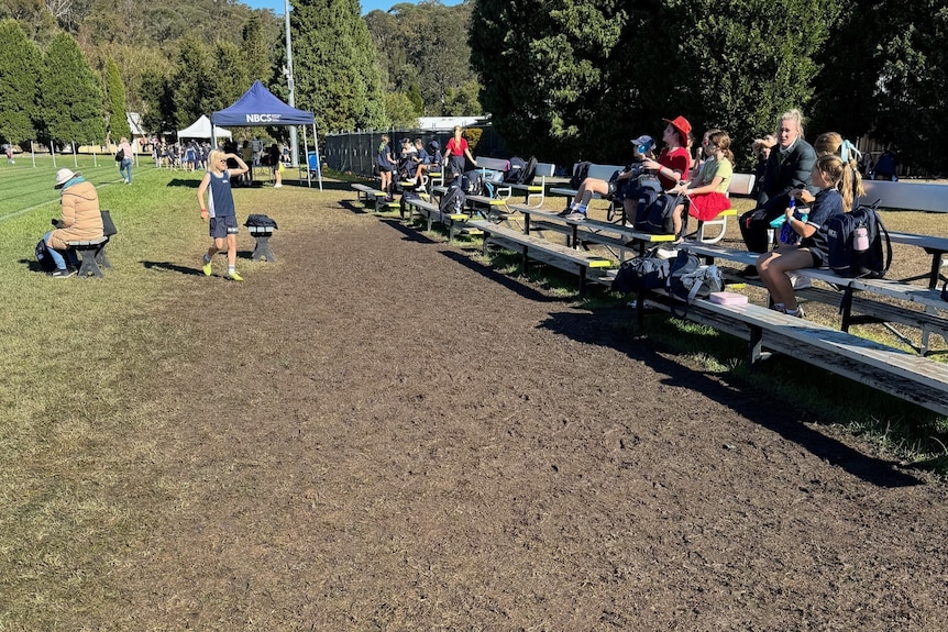 A small crowd sit on silver seats in front of muddy grass.