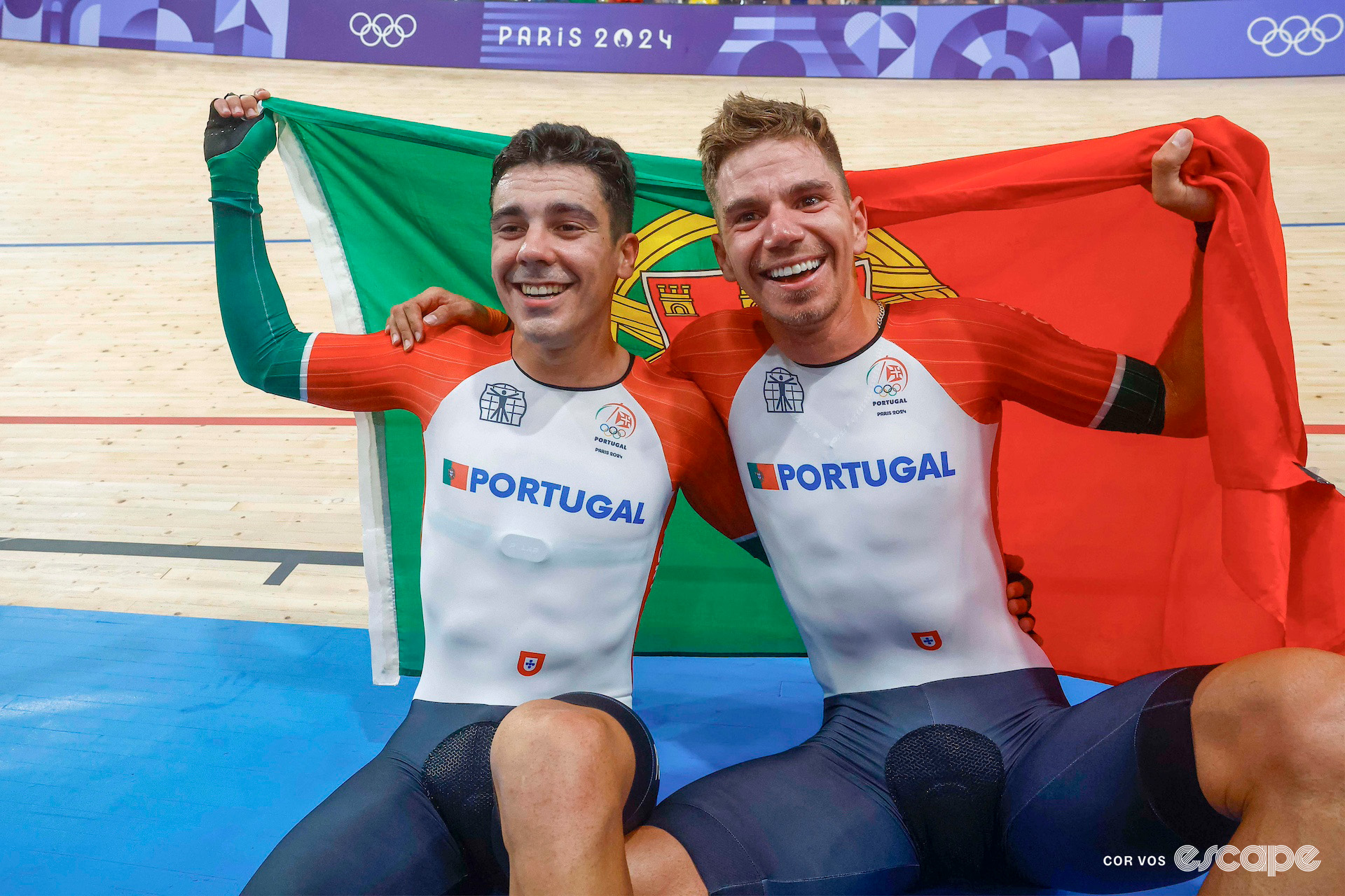 Iúri Leitão and Rui Oliveira sit on the edge of the track holding a Portugal flag behind them as they celebrate Olympic madison gold.