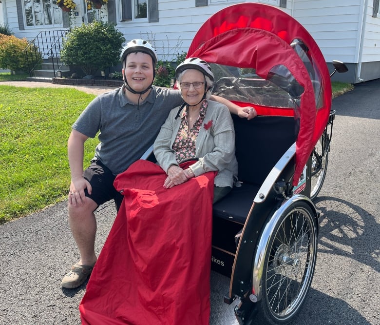 A man stands beside an older woman sitting in a tricycle with a seat attached to the front.