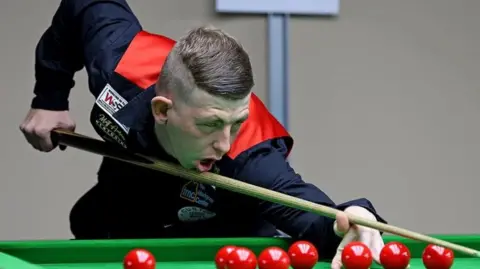 Ryan Watterson WPBSA/WDBS David Church playing snooker while wearing a red waistcoat and black shirt. Under a bright light, he is leaning over the green felt of a table, his cue at the ready, concentrating on a shot.