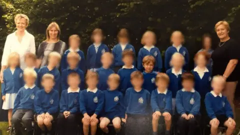 Chew Stoke Church School A class picture taken when Lando Norris was a pupil at the primary school. There are three rows of children wearing blue sweatshirts with the school logo on it. They are standing outside with trees in the background. Three female teachers stand at the back of the children smiling at the camera. All of the children's faces have been blurred except for Lando Norris.