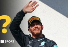 Lewis Hamilton waves while wearing a Mercedes race uniform. A question mark graphic is to the left of the image