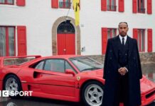 Lewis Hamilton stood alongside a Ferrari sports car while wearing a black suit and black overcoat