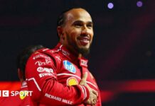 Lewis Hamilton holds his right hand across his chest while smiling at the F1 2025 season launch event. He is wearing the red Ferrari uniform 