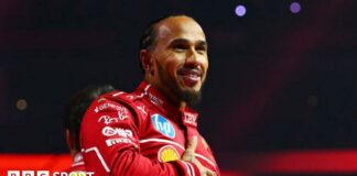 Lewis Hamilton holds his right hand across his chest while smiling at the F1 2025 season launch event. He is wearing the red Ferrari uniform 