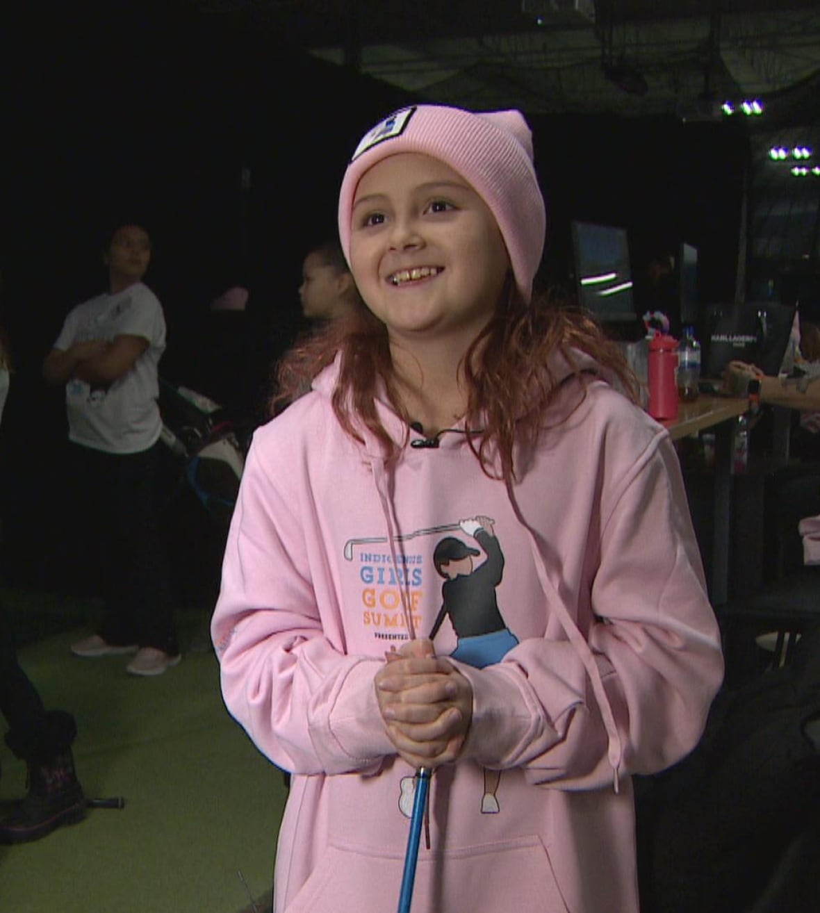 Ava smiles at the camera wearing a pink sweatshirt with a female golfer wearing a blue ribbon skirt.