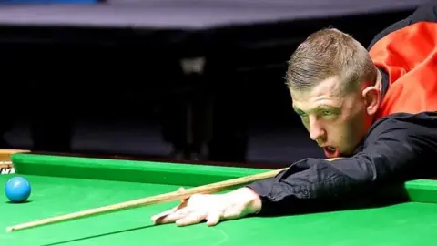 Ryan Watterson WPBSA/WDBS David Church playing snooker while wearing a red waistcoat and black shirt. Under a bright light, he is leaning over the green felt of a table, his cue at the ready, concentrating on a shot. 