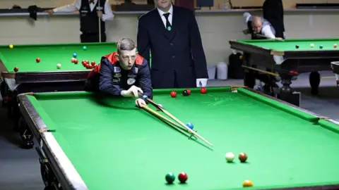 Ryan Watterson WPBSA/WDBS David Church wearing a black and red waistcoat leaning over a snooker table and taking a shot while using a rest. A referee is standing behind him. Play is under way at two other snooker tables, which can be seen behind Mr Church.