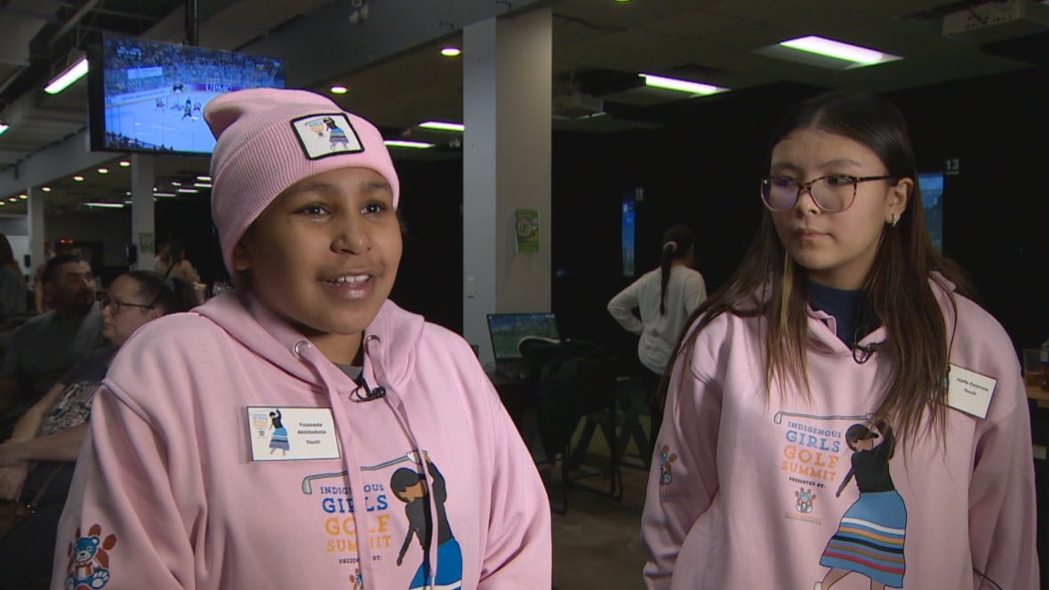 Two girls stand inside an indoor golf course wearing matching pink sweatshirts and toques.