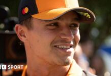 Lando Norris smiles while wearing a McLaren baseball cap with a camera filming him from behind