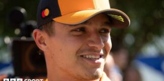 Lando Norris smiles while wearing a McLaren baseball cap with a camera filming him from behind