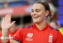 Danielle Gibson holds up her palm and is wearing a red England cricket shirt