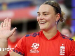 Danielle Gibson holds up her palm and is wearing a red England cricket shirt